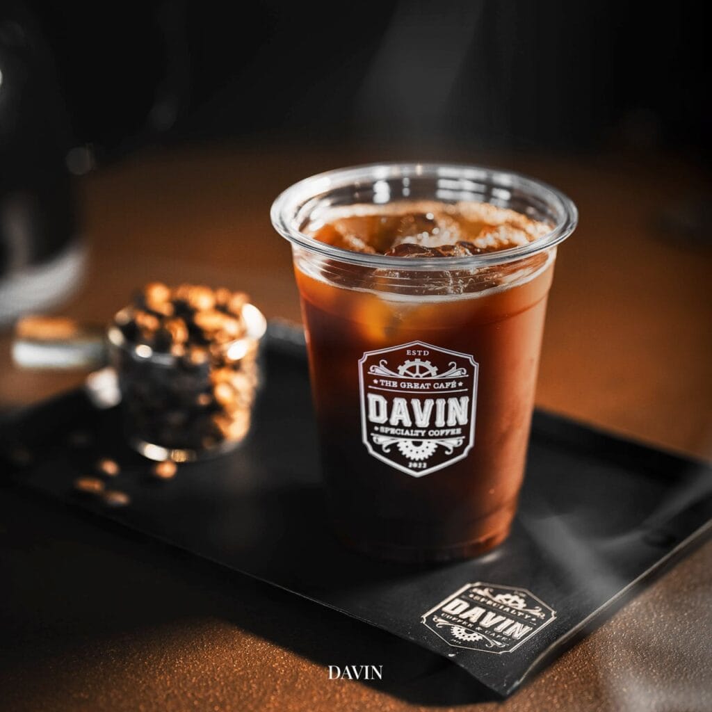 Close-up of Davin Cafe Ramintra's signature iced specialty coffee in a branded plastic cup, next to coffee beans on a black tray.