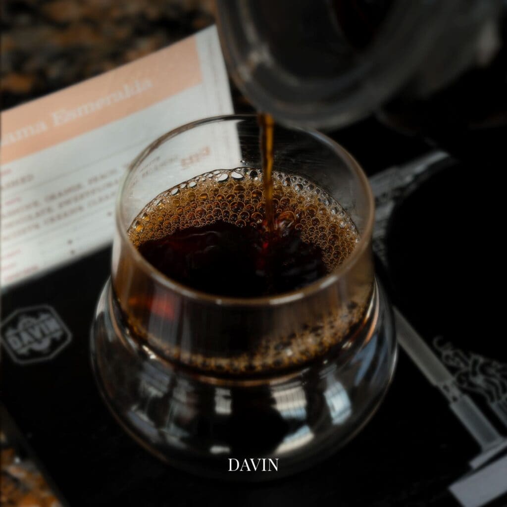 Freshly brewed specialty coffee being poured into a stemmed tasting glass, with tasting notes visible in the background.
