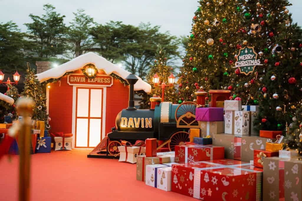 มุมถ่ายรูปคริสต์มาสสุดอลังการของ Davin Cafe ประกอบด้วยโมเดลรถไฟ "Davin Express", บ้านหลังเล็กสีแดง และกล่องของขวัญมากมาย.