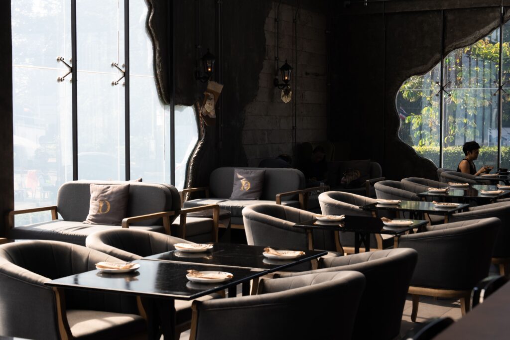 The spacious interior of a uniquely designed coffee shop featuring comfortable dining chairs, sleek tables, and large free-form windows that allow natural light to complement the dark industrial aesthetic.