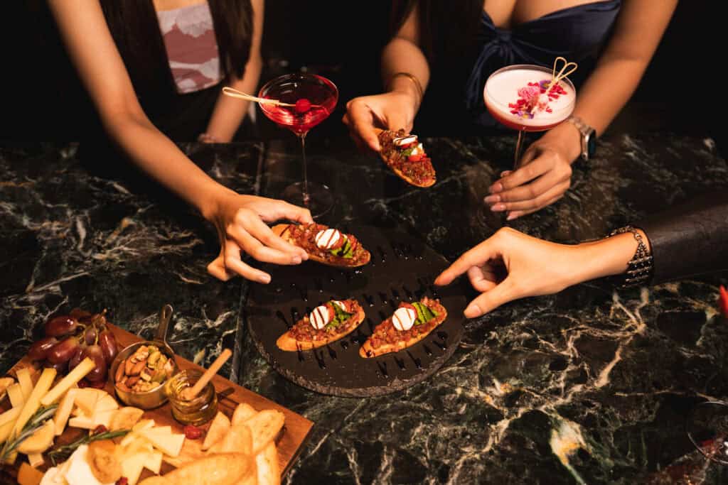 Guests sharing cocktails and small plates on a marble table at VINSENS, capturing a stylish chill bar in Bangkok for social drinks, casual conversations, flavorful bites, and relaxed nights out with a refined lounge atmosphere.